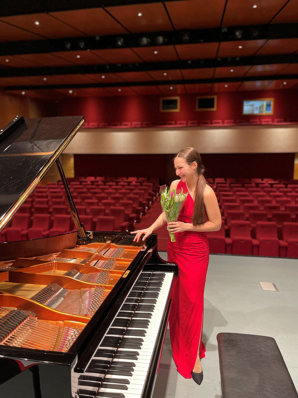 Angela Todorova in a red gown beside the piano in an empty concert hall